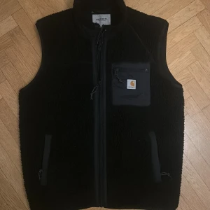 Svart fleeceväst från Carhartt - Svart fleeceväst från Carhartt med dragkedja framtill och en bröstficka med logotyp. Västen har två sidofickor med dragkedja och hög krage. Perfekt för lager-på-lager och streetwear-stil.
