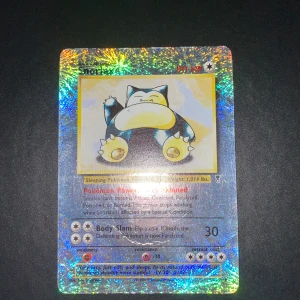 Snorlax holografiskt 64/110 Pokémonkort - Säljer ett holografiskt Snorlax Pokémonkort med glittrig yta. Kortet har en bild på Snorlax i mitten och text om dess förmågor och attacker. Baksidan har klassisk blå Pokémon-design med Pokéboll. - Skriv vid intresse 