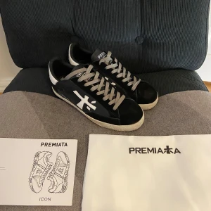 Premiata Sneaker - Säljer ett par svarta sneakers från Premiata med vit logga på sidan och grå snörning. Skorna har en platt sula i beige och detaljer i vitt på hälen. Perfekta för dig som gillar stilrena och sportiga skor.