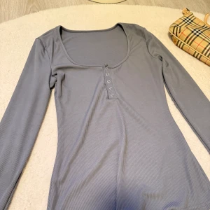 Grå långärmad ribbad topp med knappar - Snygg grå långärmad topp med ribbad struktur och knappar framtill vid halsringningen. Djup rundad urringning ger en modern look. Perfekt att styla till jeans eller kjol.