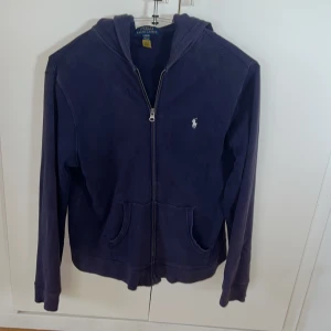 Mörkblå hoodie från Polo Ralph Lauren strl Xs  - Säljer en mörkblå hoodie tunnare variant, perfekt till vår-sommar-höst.  från Polo Ralph Lauren med dragkedja framtill och två fickor. Klassisk broderad logga på bröstet och huva. Perfekt för en avslappnad stil. Strl XS vuxen eller XL barnmodell. Nypris 1600kr inköpt på Ralph laurens hemsida