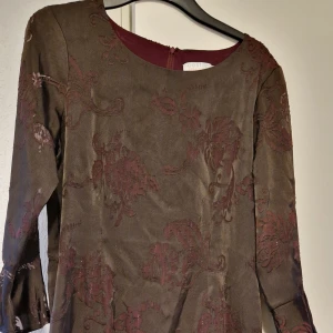 Brun klänning med blommigt mönster från Coster Copenhagen - Säljer en brun klänning från Coster Copenhagen med vinröda blommiga broderier. Klänningen har trekvartsärm med utsvängda ärmslut och rund halsringning. Dragkedja bak för enkel påklädning. Perfekt för dig som gillar unika detaljer.