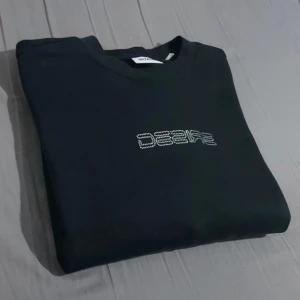 Svart sweatshirt från Weekday med strasstext - Säljer en svart sweatshirt från Weekday med texten 'DESIRE' i strasstenar på bröstet. Tröjan har rund halsringning och långa ärmar. Perfekt för en stilren och trendig look.