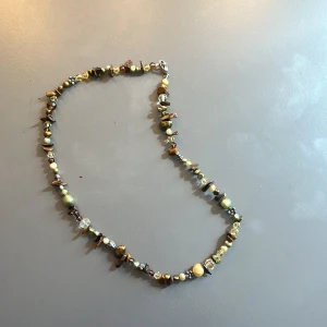 Halsband med pärlor och stenar i jordnära toner - Handgjort halsband med blandade pärlor och stenar i bruna, gula och gröna nyanser. Inslag av skimrande och transparenta pärlor samt oregelbundna stenar.