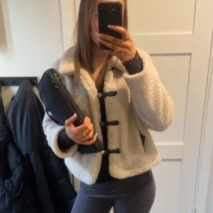 Beige teddyjacka med svarta detaljer - Säljer en beige teddyjacka med svarta knäppningar och detaljer. Jackan har en bred krage och är långärmad. Perfekt för dig som vill ha en mysig och trendig look. Passformen är normal och materialet är mjukt och fluffigt.