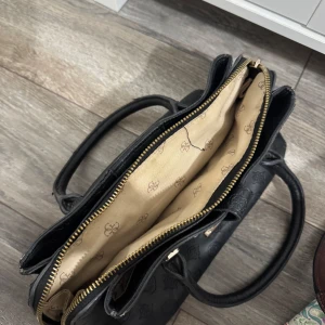 Svart handväska från Guess - Säljer en svart handväska från Guess med guldfärgade metalldetaljer och präglat mönster över hela väskan. Väskan har dubbla handtag och dragkedja upptill. Perfekt för att bära dina viktigaste saker med stil.