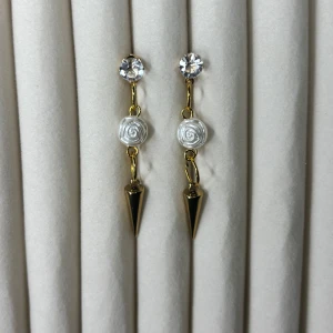 Guldiga örhängen med vita rosor och kristall - Snygga örhängen i guld med glittrande kristall upptill, en vit pärlros i mitten och en spetsig guldig detalj längst ner. Perfekta för att ge din look en unik och elegant touch.