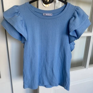 Blå ribbad topp med volangärmar  - Supersöt ljusblå topp från Zara med ribbad struktur och stora volangärmar. Perfekt för dig som vill ha något enkelt men ändå med en twist. Rund halsringning och mjukt material.