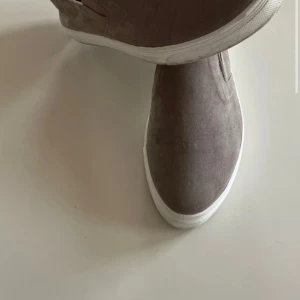 Beige lofers sneakers i mocka - Säljer ett par stilrena slip-on sneakers i beige mocka med vit platt sula. Skorna har en minimalistisk design utan snörning och är perfekta för en clean look. Passar till många olika outfits och är enkla att ta på och av.