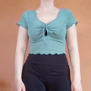Croppad topp med knyt - Blågrön croppad topp med lätt ribbat tyg.  Toppen har v-ringning och man kan knyta en rosett framtill. Kanterna på ärman och nedertill är vågade. Köpt på Sellpy men ursprungligen från Shein, mycket bra skick och använd fåtal gånger. Storlek S 💙