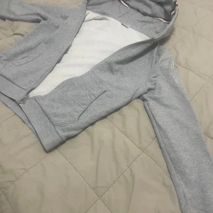 Grå hoodie från Moncler med dragkedja - Säljer en stilren grå hoodie från Moncler med dragkedja och huva. Jackan har ribbade muddar och Moncler-logga på ärmen. Insidan är vit och luvan har en snygg randig detalj. Perfekt för en avslappnad look.