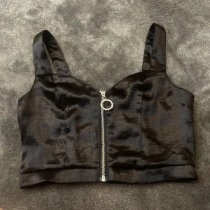 Svart croppad linnetopp med dragkedja - Snygg svart croppad linnetopp i glansigt material med breda axelband och dekorativ dragkedja framtill. Baksidan har smock för extra komfort och passform. Perfekt att styla till olika outfits!