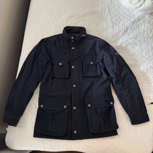 Mörkblå fodrad jacka från Hackett London - Säljer en mörkblå jacka från Hackett London i storlek M. Jackan har fyra framfickor med lock och knappar, hög krage och quiltat foder. Stängs med både dragkedja och knappar. Perfekt för kyligare dagar och har en klassisk, stilren look. Jackan är fodrad samtidigt som materialet klarar av regn. Väl använd, med visst slitage. Inhandlad på NK man i Stockholm för 5000 kr. 