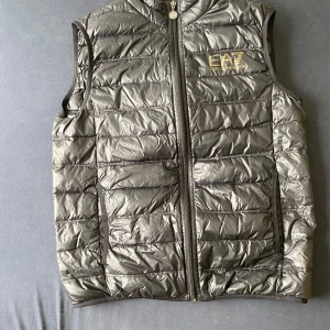 Svart dunväst från EA7 Emporio Armani - Snygg svart dunväst från EA7 Emporio Armani med guldfärgad logga på bröstet. Västen har dragkedja framtill och quiltad design. 