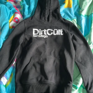 Svart hoodie från DirtCult San Diego - Säljer en svart hoodie från DirtCult San Diego med vit logga tryckt på bröstet. Tröjan har huva och en stor magficka framtill. Perfekt för dig som gillar streetwear och vill ha en enkel men snygg look. Med storlek 158/164