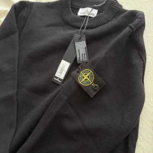 Svart stickad tröja från Stone Island - Säljer en svart stickad tröja från Stone Island med klassisk rund halsringning och ribbade muddar. Tröjan har den ikoniska Stone Island-loggan på vänster ärm. Perfekt för en stilren och avslappnad look.