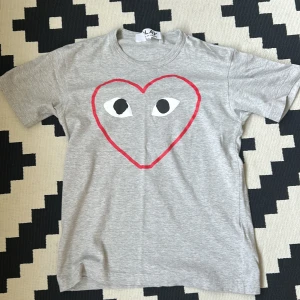 Grå t-shirt från Comme des Garçons Play - Säljer en grå t-shirt från Comme des Garçons Play med det ikoniska röda hjärtmotivet och ögon på bröstet. T-shirten är kortärmad och tillverkad i mjuk bomull. Perfekt för en stilren och trendig look.