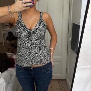 Svartvit mönstrad linnetopp - Säljer en svartvit linnetopp med smala axelband och v-ringning. Toppen har ett abstrakt mönster och en liten knytning framtill. Perfekt för varma dagar och enkel att matcha med jeans eller kjol.