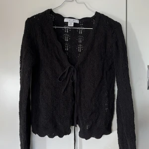 Svart stickad kofta från Amisu - Säljer en svart stickad kofta från Amisu i storlek XS. Koftan har lång ärm, knytning framtill och ett fint hålmönster i tyget. Perfekt att slänga över en topp för en snygg lager-på-lager-look.