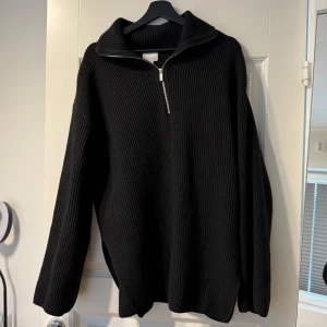 Svart stickad tröja med dragkedja - Svart stickad tröja med halv dragkedja framtill och hög krage. Tröjan har lång ärm och en avslappnad passform, perfekt för kyligare dagar.