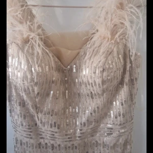 Champagnefärgad långklänning med paljetter och fjädrar - Säljer en elegant långklänning i champagnefärg med smala axelband dekorerade med fjädrar. Klänningen är täckt av glittriga paljetter och har en v-ringad urringning. Perfekt för bal eller festliga tillfällen.