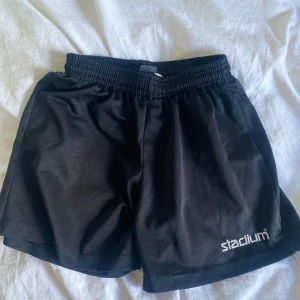 Svarta träningsshorts från Stadium - Svarta träningsshorts från Stadium med elastisk midja och logga på ena benet. Perfekta för sport eller gympa. Lätta och bekväma med normal passform.