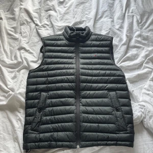 Svart dunväst från Tommy Hilfiger - Snygg svart dunväst från Tommy Hilfiger med horisontella sömmar och dragkedja framtill. Västen har ståkrage och två fickor med och enkel att matcha.