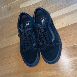 Svarta Vans Old Skool sneakers - Klassiska svarta Vans Old Skool sneakers med svart sula och snörning. Skorna har den ikoniska sidoremsan i svart och är tillverkade i mocka och syntet. Perfekta för en streetwear-look och passar till det mesta.