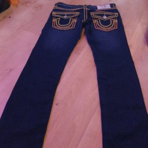 Mörkblå jeansbyxor med kontrastsömmar från True Religion - Säljer ett par mörkblå jeansbyxor från True Religion med tydliga gula kontrastsömmar och stora fickor bak med dekorativa knappar. Klassisk femficksmodell och raka ben. Perfekta för dig som gillar streetwear och vill sticka ut.