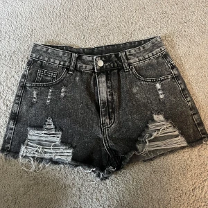 Gråa jeansshorts med slitningar - Säljer ett par svarta jeansshorts med slitningar nertill. Shortsen har fyra fickor, bälteshällor och knappstängning. Perfekta för en cool och avslappnad stil.