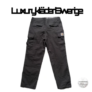 Carhartt cargobyxor Svarta - Svarta cargobyxor från Carhartt med flera fickor på sidorna och bak. Byxorna har raka ben och är tillverkade i en slitstark bomulls- och polyesterblandning. Klassisk Carhartt-logga på ena benfickan och metallknapp med varumärkesprägling.storlek M-L