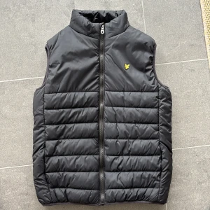 Svart dunväst från Lyle & Scott  - Säljer en svart dunväst från Lyle & Scott Junior med gul logga på bröstet. Västen har ståkrage och dragkedja framtill. Perfekt för lager-på-lager och håller dig varm under kyliga dagar. Använd 1 gång. Pris kan diskuteras!