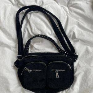 Svart axelväska från Noella - Säljer en svart axelväska från Noella i pälsimitation. Väskan har två framfickor med dragkedja och en justerbar axelrem med vita prickar. Perfekt för att bära det viktigaste och ge en cool touch till din outfit. Har inte använt den alls och kan sälja den för mindre summa också💞💞