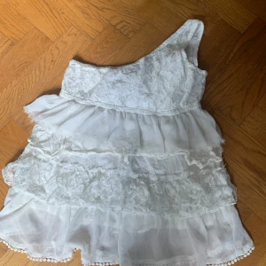 Vit spets blus med volanger från Zheno/Elios - Söt vit blus med spetsdetaljer och volanger. Blusen har oneshoulder-modell och flera lager med spets och tyll. Perfekt för sommarens tillfällen och har en romantisk känsla. Skulle säga att den passar folk som har mindre storlek än 40 också.