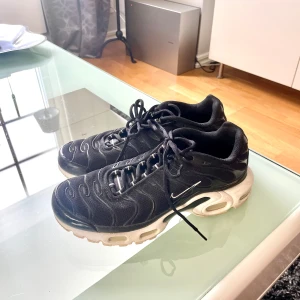 Svarta Nike Air Max TN sneakers - Säljer ett par svarta Nike Air Max TN sneakers med vit sula och klassisk Tn-detalj på hälen. Skorna har snörning, rund tå och synliga vågmönster på ovandelen. Perfekta för dig som gillar sportig och stilren look.