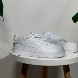 Nike Air Force 1 vita sneakers - Klassiska helvita Nike Air Force 1 sneakers med rund tå och platt sula. Skorna har snörning och är tillverkade i syntetmaterial med diskret Nike-logga på sidan. Perfekta för en clean och stilren look. Finns i storlek 41 & 45.