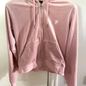 Rosa velour hoodie från Juicy Couture - Säljer en ljusrosa hoodie i mjuk velour från Juicy Couture. Tröjan har dragkedja, huva med snören och en broderad logga på bröstet. Praktisk känguruficka framtill och croppad passform. Perfekt för en avslappnad och trendig look!