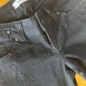 Y2K Svarta jeans med låg midja med strasstenar från Vero Moda  - Snygga svarta lågmidjade jeans från Vero Moda  med raka ben och dekorativa strasstenar i ett mönster längs ena benet. Klassisk femficksmodell med knappar och nitar. Perfekta för dig som vill sticka ut lite extra.                                                         Storlek är oklart men passar som XS-S.  Midjemått tvärs över är 34cm och innerbendlängden är 80cm.