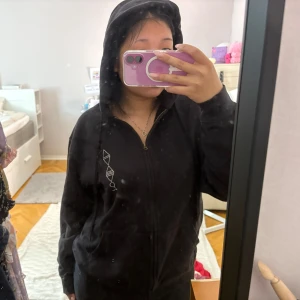 Svart hoodie med tryck från Ghost - Säljer en svart hoodie med dragkedja från Ghost. Framsidan har en vit geometrisk design och baksidan har ett stort tryck med bandets logga och motiv. Tröjan har huva och fickor framtill, perfekt för dig som gillar bandmerch.