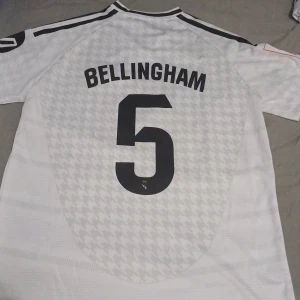 Real Madrid fotbollströja Bellingham 5 - Vit Real Madrid matchtröja med svarta detaljer, Bellingham och nummer 5 på ryggen. Adidas-logga och klubbmärke på bröstet, samt sponsortryck 'Emirates Fly Better' på framsidan. Kortärmad modell med subtilt mönster i tyget.