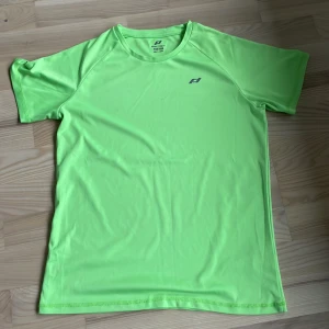 Grön tränings-t-shirt från Pro Touch - Säljer en neongrön tränings-t-shirt från Pro Touch i storlek 158/164. T-shirten är tillverkad i ett lätt och snabbtorkande syntetmaterial, perfekt för träning eller sport. Klassisk passform med korta ärmar och rund hals.
