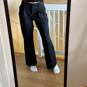 Svarta kostymbyxor med pressveck - Snygga svarta kostymbyxor med pressveck och vida ben. Byxorna har hög midja, hällor för bälte och fickor fram. Perfekta för en clean och stilren look. Materialet känns mjukt och har en lätt struktur.