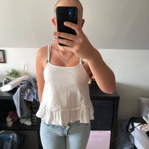 Vit linnetopp med volang från SRNHLT - Supersöt vit linnetopp från SRNHLT(Ullared) med tunna axelband och volang nertill. Toppen är luftig och har en avslappnad passform, perfekt för varma dagar. Enkel och stilren design som är lätt att matcha med jeans eller shorts.