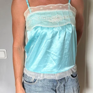 Turkos satin linnetopp med spets - Supersöt ljusblå linnetopp i satin med tunna axelband och vit spets upptill och nedtill. Toppen har broderade detaljer och en romantsik passform.