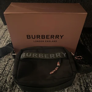 Svart axelväska från Burberry - Snygg svart axelväska från Burberry med tydlig logga på remmen och dragkedja. Väskan har ett stilrent och sportigt uttryck, justerbar axelrem och invändiga kortfack. Materialet är slitstark syntet och detaljer i skinn. Perfekt för dig som vill ha en clean och lyxig look.