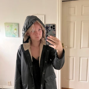 Svart regnjacka med teddyfoder - Svart regnjacka med mysigt beige teddyfoder och huva. Jackan har stora fickor framtill, dragkedja och tryckknappar. Perfekt för ruggiga dagar när du vill hålla dig både torr och varm. Snygg och enkel design som funkar till det mesta. Den är förstås lite liten på mig (jag är 158cm och 18 år)