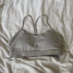 Grå sportig linnetopp från Xshapes - Snygg grå linnetopp från Xshapes med smala axelband och brottarrygg. Perfekt för träning eller chill dagar. Mjuk och stretchig syntetblandning som sitter skönt mot kroppen. Diskret logga framtill på resåren.