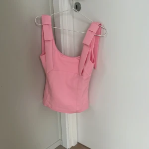 Rosa linnetopp med rosettband i storlek xs - Supersöt ljusrosa linnetopp med breda axelband som knyts i stora rosetter. Toppen har en figurnära passform och ärmlös design, perfekt för varma dagar. Materialet känns mjukt och lätt mot huden. En riktig statement-piece för dig som gillar färg och detaljer. Den är väldigt stor i storleken och känns inte som en xs så en person som oftast brukar ha S skulle denna passa perfekt på💗