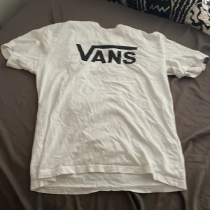 Vit Vans t-shirt med logga - Vit t-shirt från Vans med stor svart logga på bröstet. Klassisk passform med rund hals och korta ärmar. Perfekt basic-plagg för dig som gillar streetwear och enkel stil.