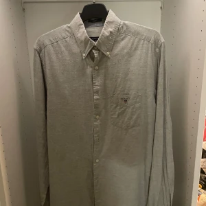 Grå skjorta från Gant - Snygg grå skjorta från Gant i bomull med klassisk krage och knäppning framtill. Bröstficka med broderad Gant-logga och sköld. Perfekt till jeans eller chinos för en clean look. Aldrig använd